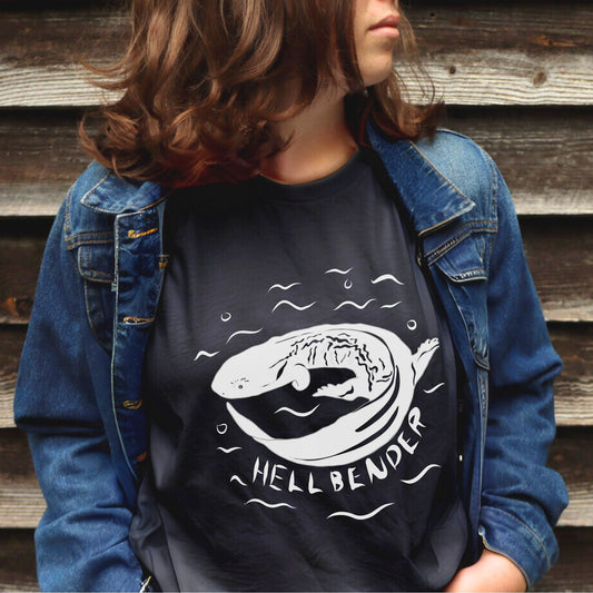 Woman in black Hellbender t-shirt with a jean jacket in front of wooden siding. White graphic and text design on the comfort colors tee.