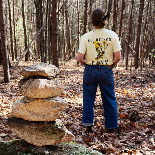 Woman standing next to a rock stack in Butter "Goldfinch Love" comfort colors t-shirt featuring vintage style graphic of two goldfinches on a thistle brance between bold text.