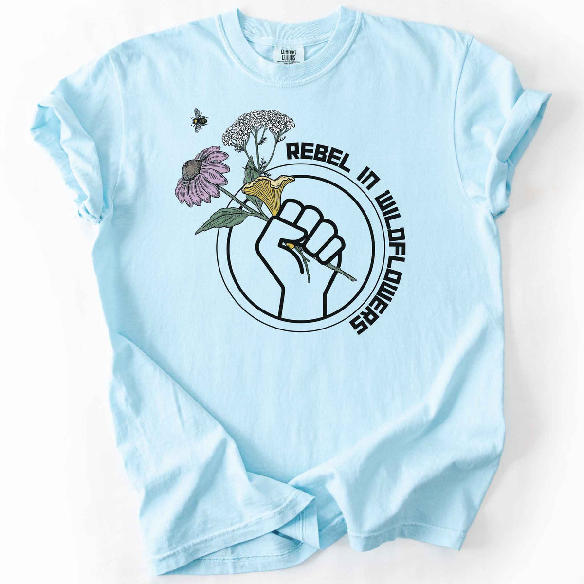 Chambray "Rebel in Wildflowers" t-shirt on Comfort Colors tee, featuring a raised fist holding coneflower, yarrow, and a mushroom with a bumblebee in a circular badge design. Gardener and native plant gift shirt for wildflower and pollinator conservation fans.