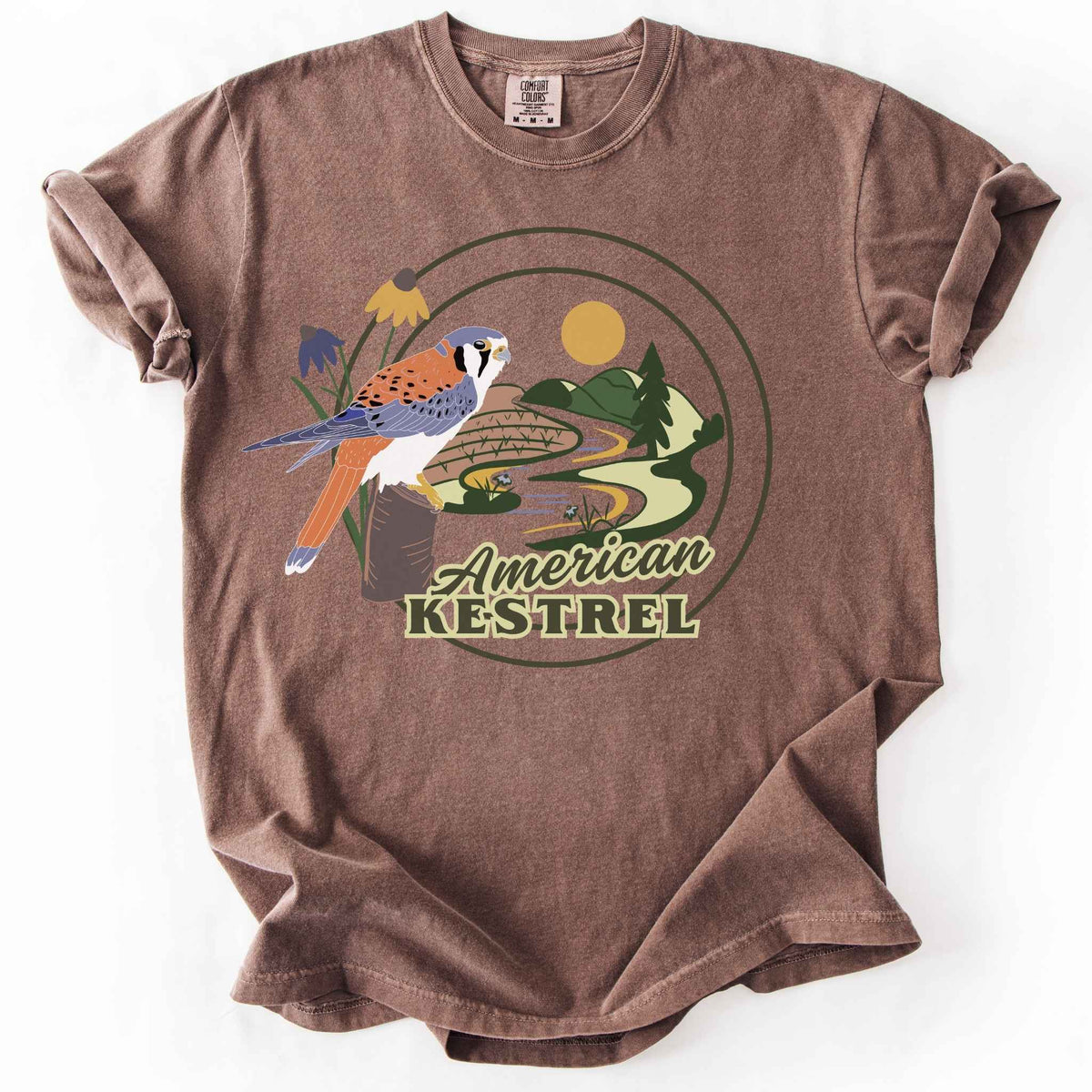 Espresso "American Kestrel" t-shirt on Comfort Colors tee, featuring a colorful illustrated kestrel perched among wildflowers with a farmland and river landscape in a circular badge design. Birdwatcher and raptor lover gift shirt for wildlife and nature enthusiasts.
