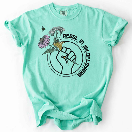 Island Reef "Rebel in Wildflowers" t-shirt on Comfort Colors tee, featuring a raised fist holding coneflower, yarrow, and a mushroom with a bumblebee in a circular badge design. Gardener and native plant gift shirt for wildflower and pollinator conservation fans.
