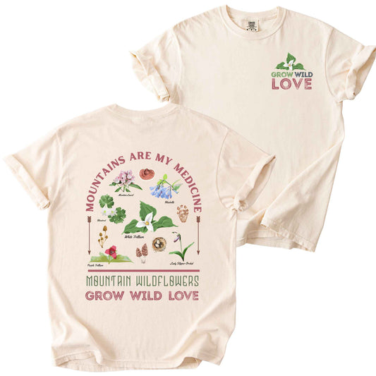 Ivory "Mountains Are My Medicine, Mountain Wildflowers, Grow Wild Love" t-shirt on Comfort Colors tee, featuring a small front chest logo and a full illustrated back print of labeled wildflowers and mushrooms including Trillium, Bluebells, and Lady Slipper Orchid. Botanical gift shirt for hikers and wildflower enthusiasts.