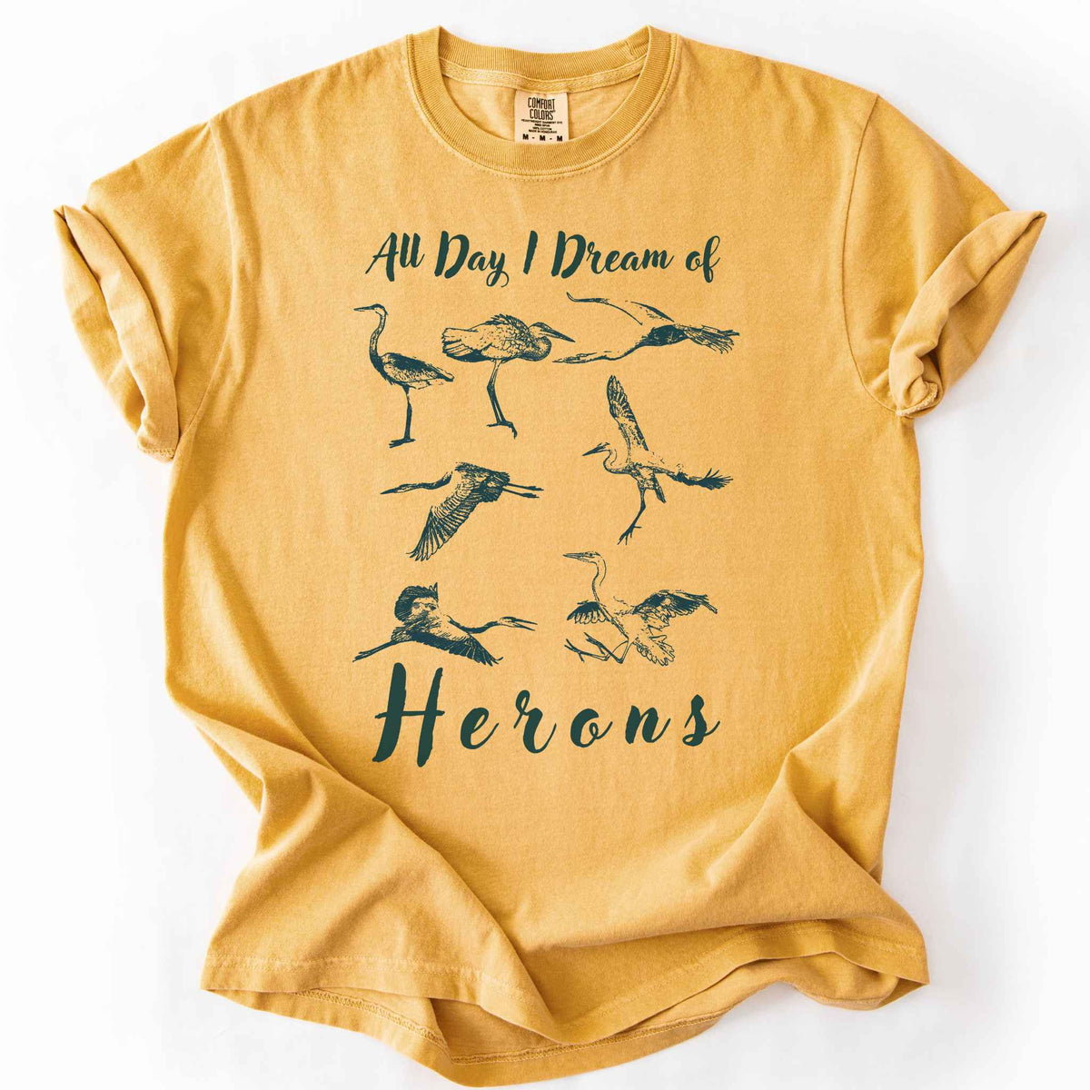 Mustard "All Day I Dream of Herons" t-shirt on Comfort Colors tee, featuring seven herons in various poses including standing, flying, and hunting. Birdwatcher and wading bird lover gift shirt for wildlife and nature enthusiasts.