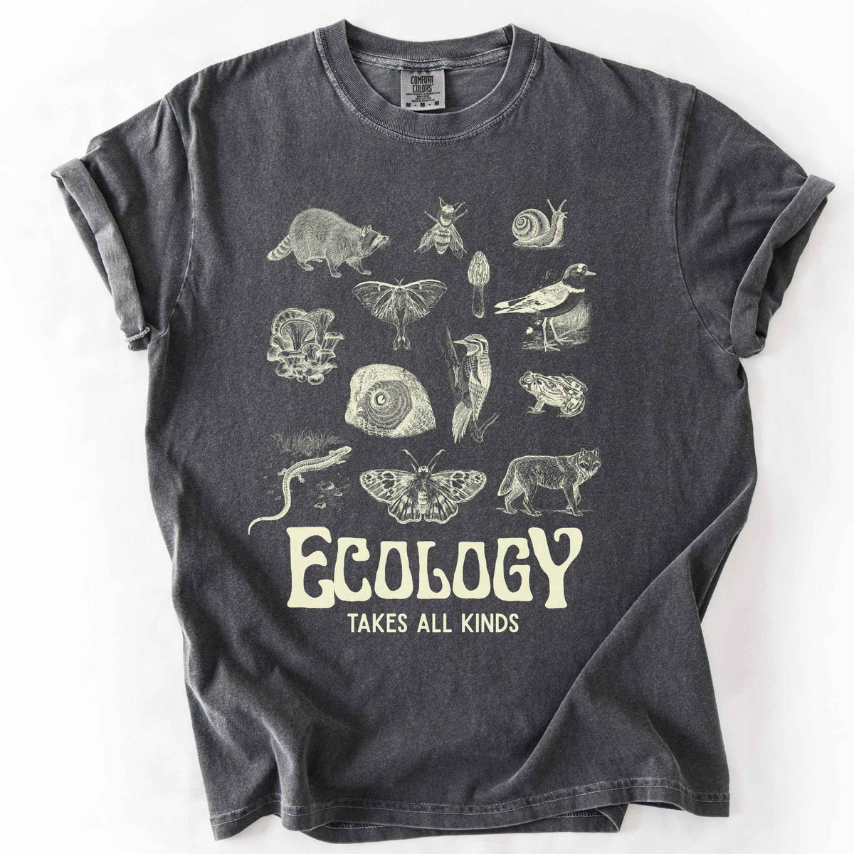 Pepper "Ecology Takes All Kinds" t-shirt on Comfort Colors tee, featuring a grid of raccoons, owls, wolves, salamanders, mushrooms, moths, and other wildlife in cream ink. Nature lover and biology gift shirt for ecologists and wildlife enthusiasts.
