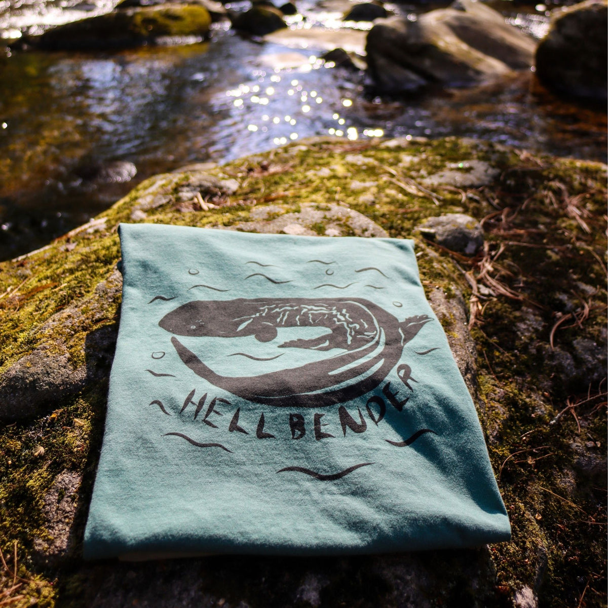 Bay Hellbender t-shirt folded and lying on a mossy rock next to a stream.