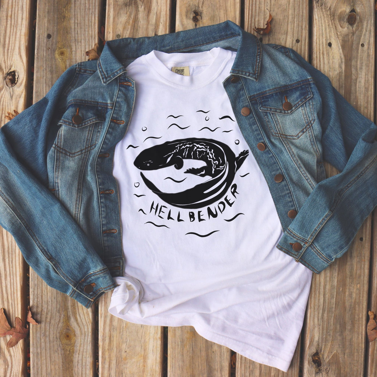 White Hellbender t-shirt in a jean jacket lying on wooden decking. Black graphic and text on comfort colors tee.