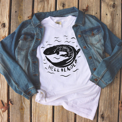White Hellbender t-shirt in a jean jacket lying on wooden decking. Black graphic and text on comfort colors tee.