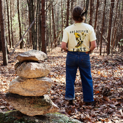 Woman standing next to a rock stack in Butter "Goldfinch Love" comfort colors t-shirt featuring vintage style graphic of two goldfinches on a thistle brance between bold text. 
