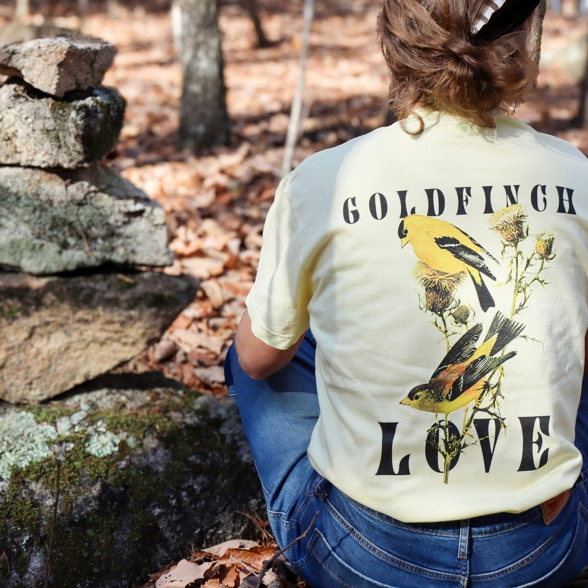 Woman sitting next to a rock stack in Butter "Goldfinch Love" comfort colors t-shirt featuring vintage style graphic of two goldfinches on a thistle brance between bold text. 
