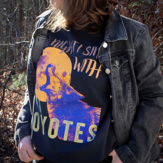 Woman wearing "All night I sing with coyotes" comfort colors t-shirt in a jean jacket in a forest setting.