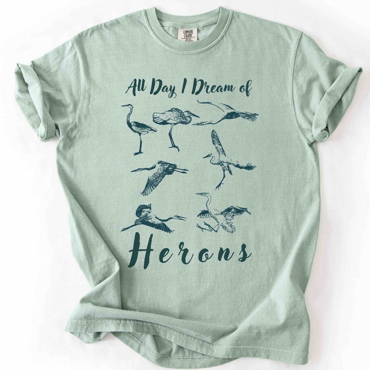 Bay "All Day I Dream of Herons" t-shirt on Comfort Colors tee, featuring seven herons in various poses including standing, flying, and hunting. Birdwatcher and wading bird lover gift shirt for wildlife and nature enthusiasts.