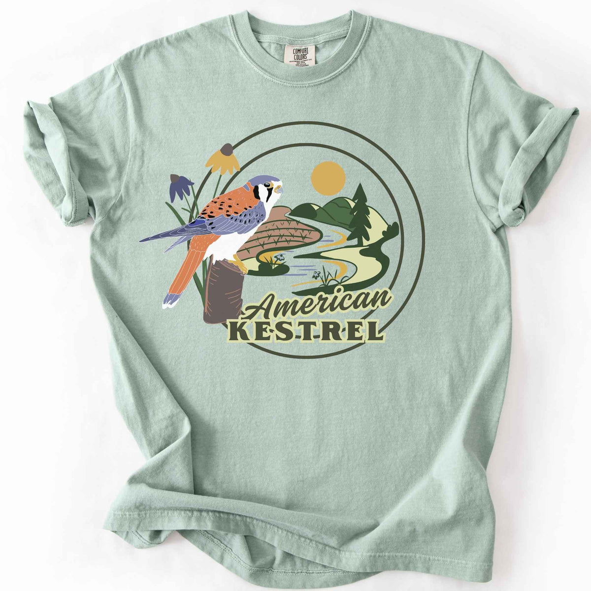 Bay "American Kestrel" t-shirt on Comfort Colors tee, featuring a colorful illustrated kestrel perched among wildflowers with a farmland and river landscape in a circular badge design. Birdwatcher and raptor lover gift shirt for wildlife and nature enthusiasts.