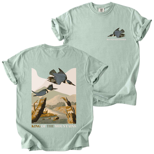 Bay "Belted Kingfisher, King of the Mountains" t-shirt on Comfort Colors tee, featuring a small front chest logo and a full illustrated back print of kingfishers over a river landscape in retro poster style. Birdwatcher gift shirt for wildlife and nature enthusiasts.