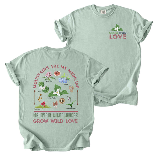 Bay "Mountains Are My Medicine, Mountain Wildflowers, Grow Wild Love" t-shirt on Comfort Colors tee, featuring a small front chest logo and a full illustrated back print of labeled wildflowers and mushrooms including Trillium, Bluebells, and Lady Slipper Orchid. Botanical gift shirt for hikers and wildflower enthusiasts.