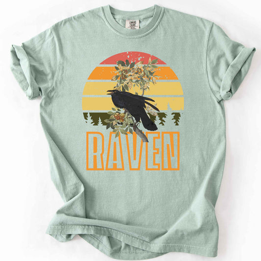 Bay "Raven" t-shirt on Comfort Colors tee, featuring a raven perched on a branch against a retro sunset sunburst design with bold orange text. Bird lover and wildlife gift shirt for corvid and nature enthusiasts.