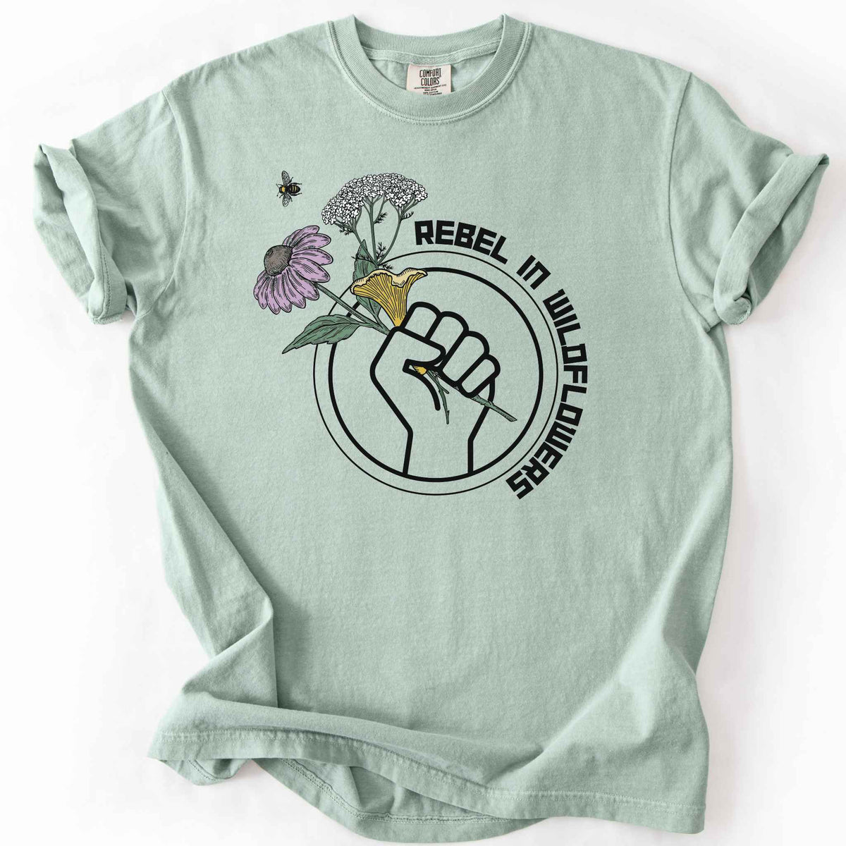 Bay "Rebel in Wildflowers" t-shirt on Comfort Colors tee, featuring a raised fist holding coneflower, yarrow, and a mushroom with a bumblebee in a circular badge design. Gardener and native plant gift shirt for wildflower and pollinator conservation fans.