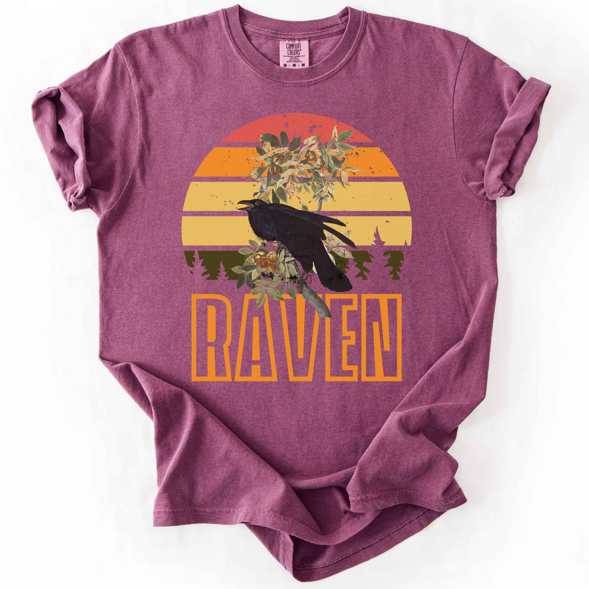 Berry "Raven" t-shirt on Comfort Colors tee, featuring a raven perched on a branch against a retro sunset sunburst design with bold orange text. Bird lover and wildlife gift shirt for corvid and nature enthusiasts.