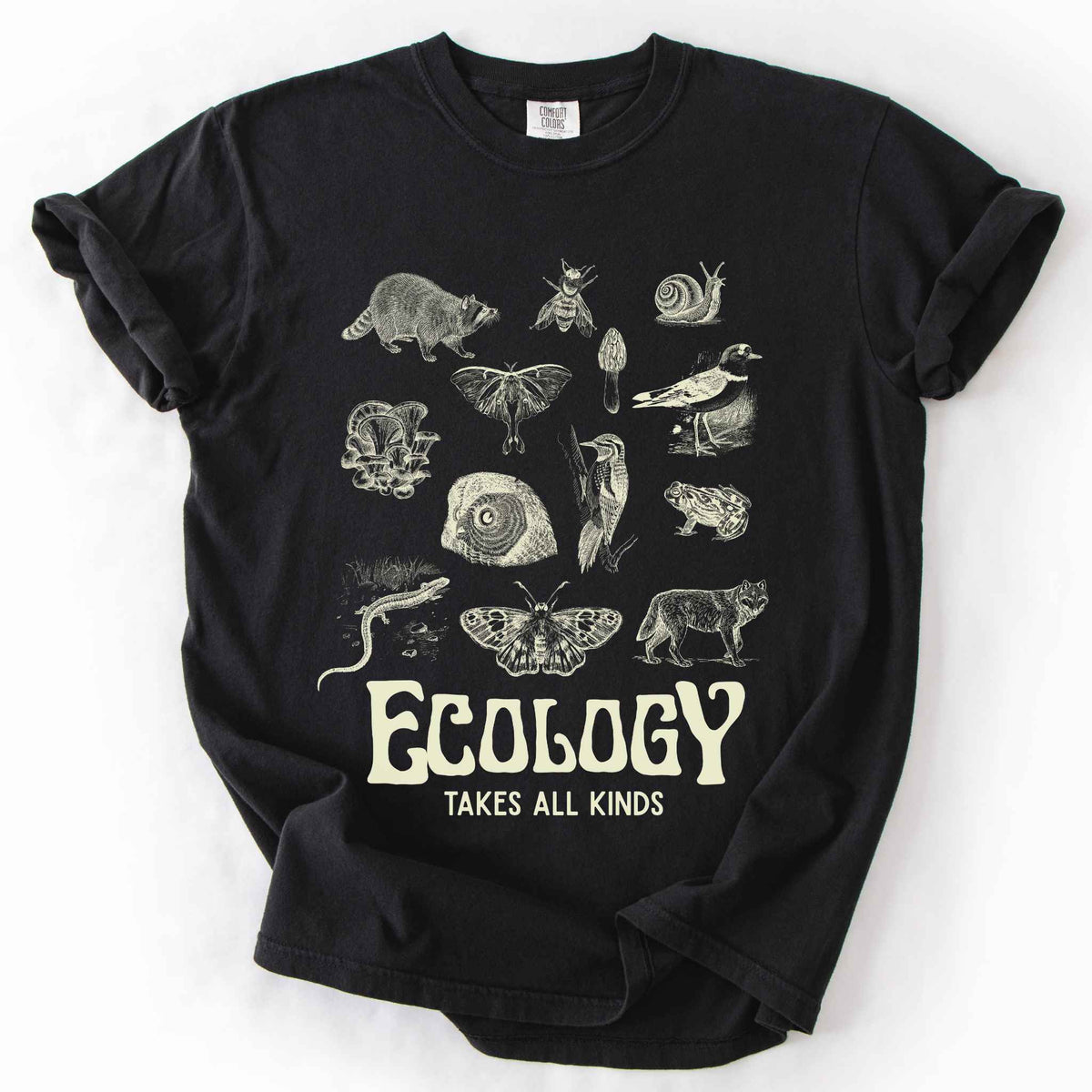 Black "Ecology Takes All Kinds" t-shirt on Comfort Colors tee, featuring a grid of raccoons, owls, wolves, salamanders, mushrooms, moths, and other wildlife in cream ink. Nature lover and biology gift shirt for ecologists and wildlife enthusiasts.