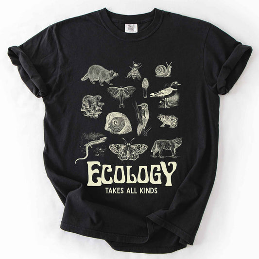 Black "Ecology Takes All Kinds" t-shirt on Comfort Colors tee, featuring a grid of raccoons, owls, wolves, salamanders, mushrooms, moths, and other wildlife in cream ink. Nature lover and biology gift shirt for ecologists and wildlife enthusiasts.