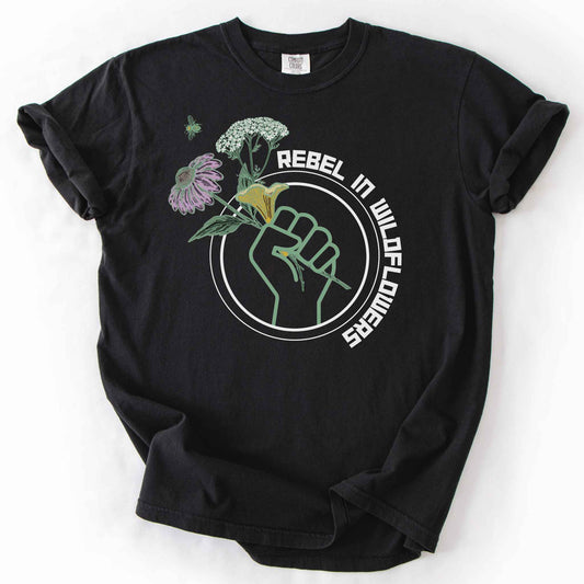 Black "Rebel in Wildflowers" t-shirt on Comfort Colors tee, featuring a raised fist holding coneflower, yarrow, and a mushroom with a bumblebee in a circular badge design. Gardener and native plant gift shirt for wildflower and pollinator conservation fans.