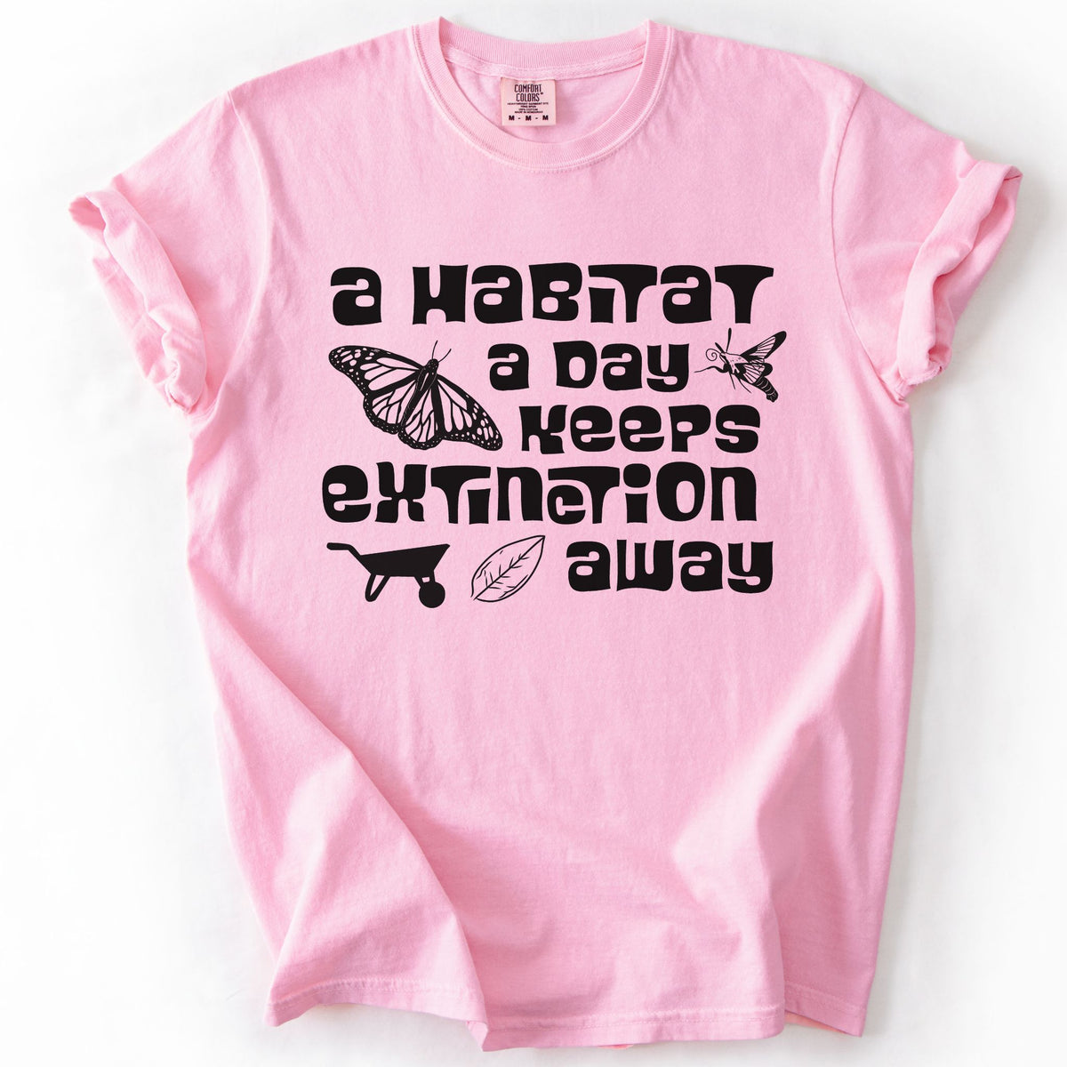 Blossom "A Habitat a Day Keeps Extinction Away" t-shirt on Comfort Colors tee, featuring a monarch butterfly, clearwing moth, wheelbarrow, and leaf icon with bold black lettering. Gardener and conservation gift shirt for native plant and pollinator enthusiasts.