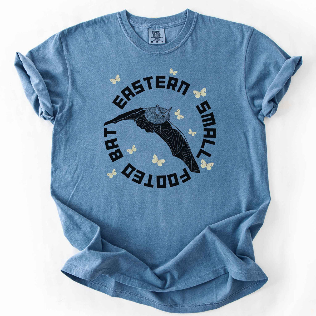 Blue Jean "Eastern Small Footed Bat" t-shirt on Comfort Colors tee, featuring a black illustration of a flying bat surrounded by pale moths in a circular badge design. Bat lover and wildlife gift shirt for nocturnal animal and nature enthusiasts.