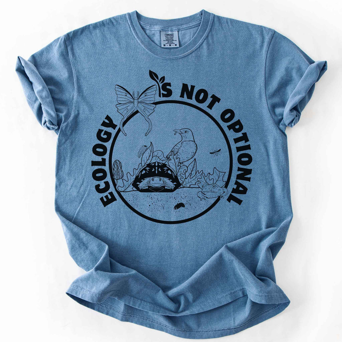 Blue Jean "Ecology Is Not Optional" t-shirt on Comfort Colors tee, featuring a black circle badge with a box turtle, bird, luna moth, frog, and mushrooms in an ecosystem scene. Nature lover and biology gift shirt for ecologists and conservation enthusiasts.