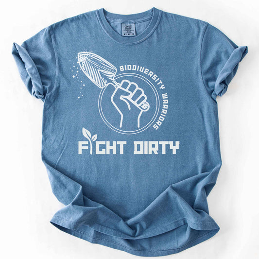 Blue jean "Biodiversity Warriors, Fight Dirty" t-shirt on Comfort Colors tee, featuring a raised fist gripping a garden trowel in a circular badge design. Gardener and conservation lover gift shirt for native plant and biodiversity enthusiasts.