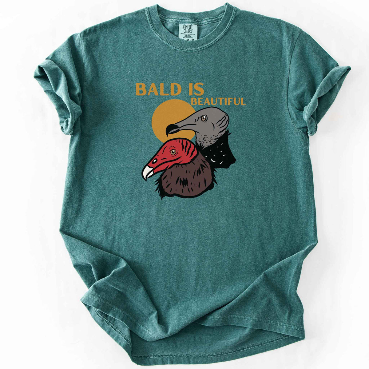 Blue spruce "Bald Is Beautiful" t-shirt on Comfort Colors tee, featuring illustrated turkey vulture and black vulture heads with orange retro sun and orange lettering. Funny birdwatcher gift shirt for vulture and scavenger lovers.