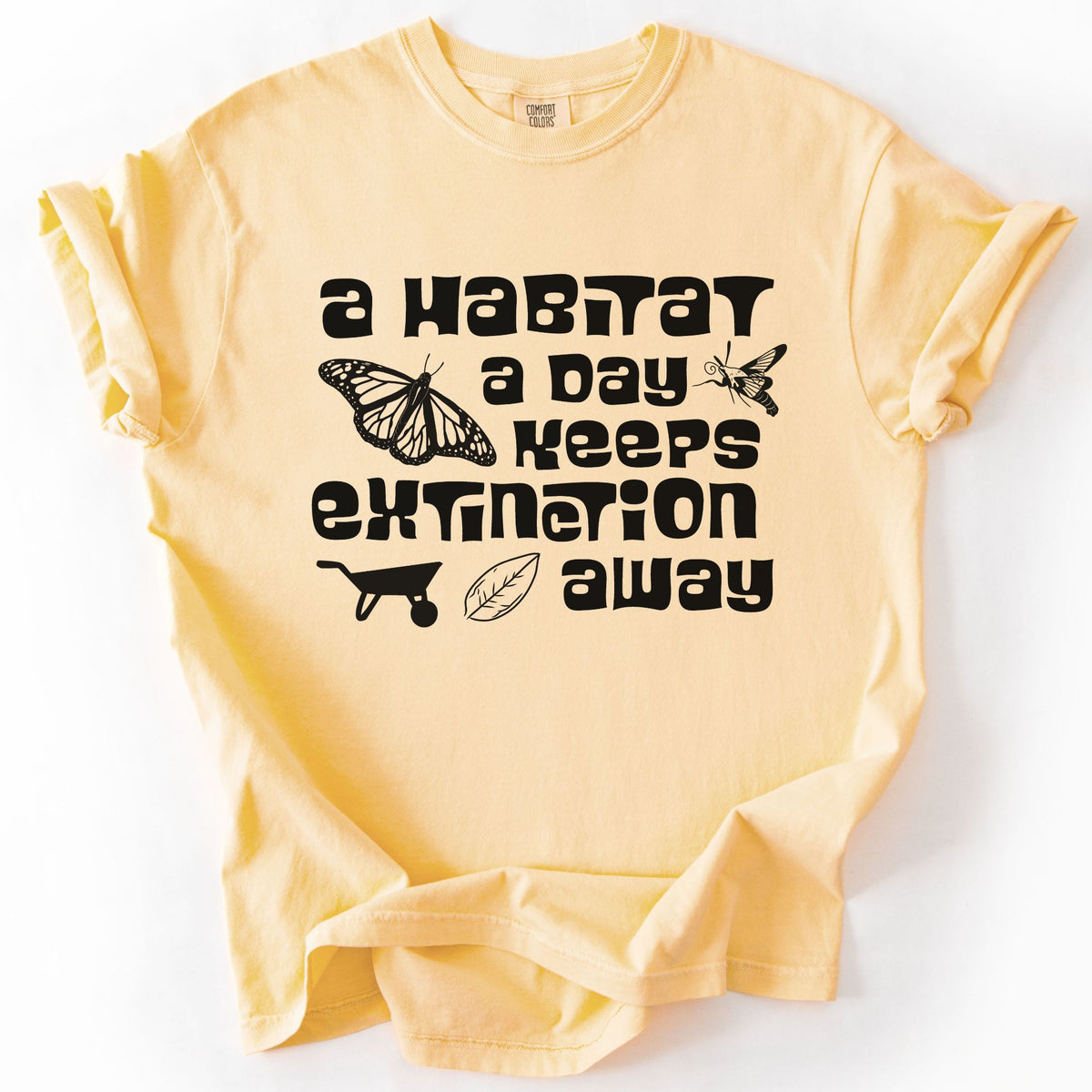 Butter "A Habitat a Day Keeps Extinction Away" t-shirt on Comfort Colors tee, featuring a monarch butterfly, clearwing moth, wheelbarrow, and leaf icon with bold black lettering. Gardener and conservation gift shirt for native plant and pollinator enthusiasts.