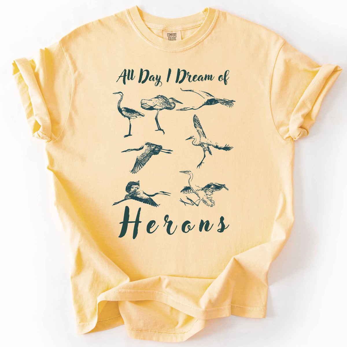 Butter "All Day I Dream of Herons" t-shirt on Comfort Colors tee, featuring seven herons in various poses including standing, flying, and hunting. Birdwatcher and wading bird lover gift shirt for wildlife and nature enthusiasts.