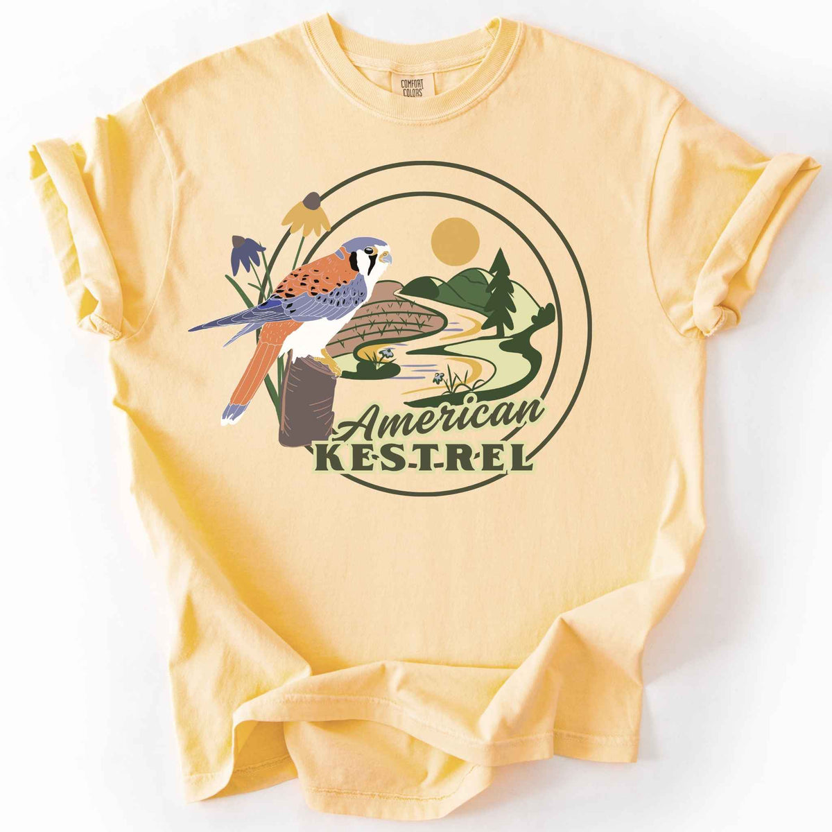Butter "American Kestrel" t-shirt on Comfort Colors tee, featuring a colorful illustrated kestrel perched among wildflowers with a farmland and river landscape in a circular badge design. Birdwatcher and raptor lover gift shirt for wildlife and nature enthusiasts.