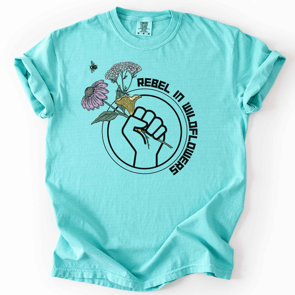 Chalky mint "Rebel in Wildflowers" t-shirt on Comfort Colors tee, featuring a raised fist holding coneflower, yarrow, and a mushroom with a bumblebee in a circular badge design. Gardener and native plant gift shirt for wildflower and pollinator conservation fans.