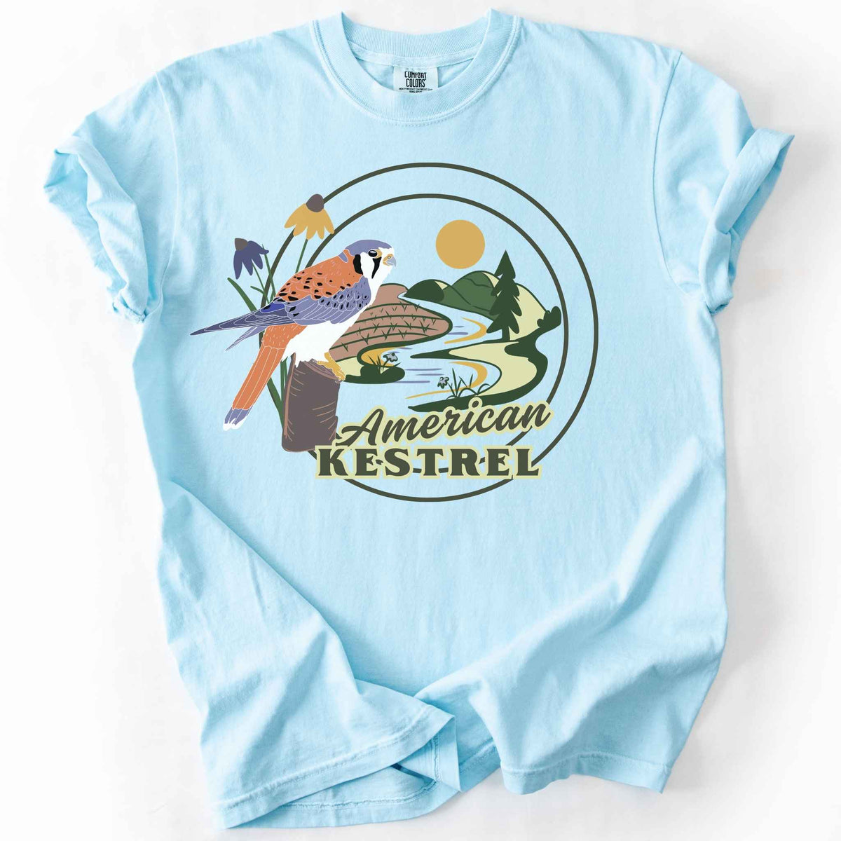 Chambray "American Kestrel" t-shirt on Comfort Colors tee, featuring a colorful illustrated kestrel perched among wildflowers with a farmland and river landscape in a circular badge design. Birdwatcher and raptor lover gift shirt for wildlife and nature enthusiasts.