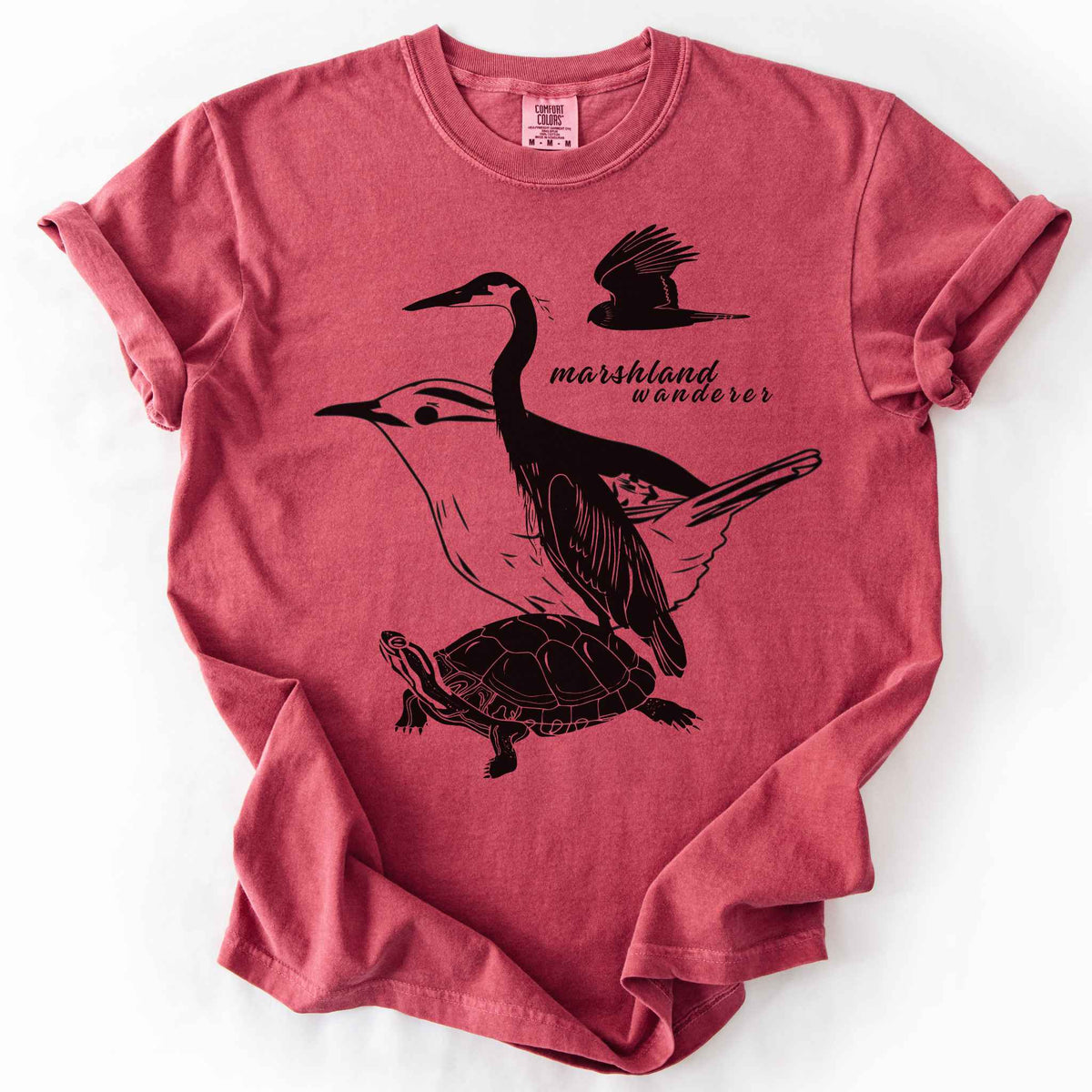 Crimson "Marshland Wanderer" t-shirt on Comfort Colors tee, featuring a surreal black line art design blending a heron, marsh wren, turtle, and marsh harrier into one flowing wetland scene. Nature lover and wetland conservation gift shirt for birdwatching and wildlife enthusiasts.