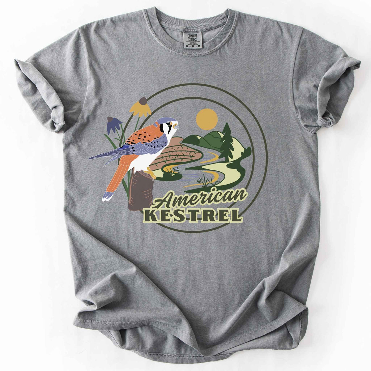 Grey "American Kestrel" t-shirt on Comfort Colors tee, featuring a colorful illustrated kestrel perched among wildflowers with a farmland and river landscape in a circular badge design. Birdwatcher and raptor lover gift shirt for wildlife and nature enthusiasts.