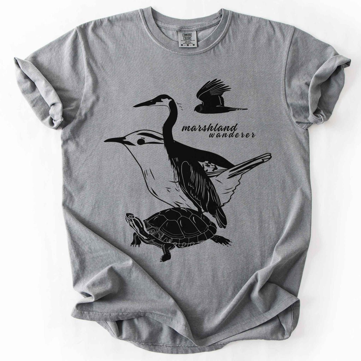 Grey "Marshland Wanderer" t-shirt on Comfort Colors tee, featuring a surreal black line art design blending a heron, marsh wren, turtle, and marsh harrier into one flowing wetland scene. Nature lover and wetland conservation gift shirt for birdwatching and wildlife enthusiasts.