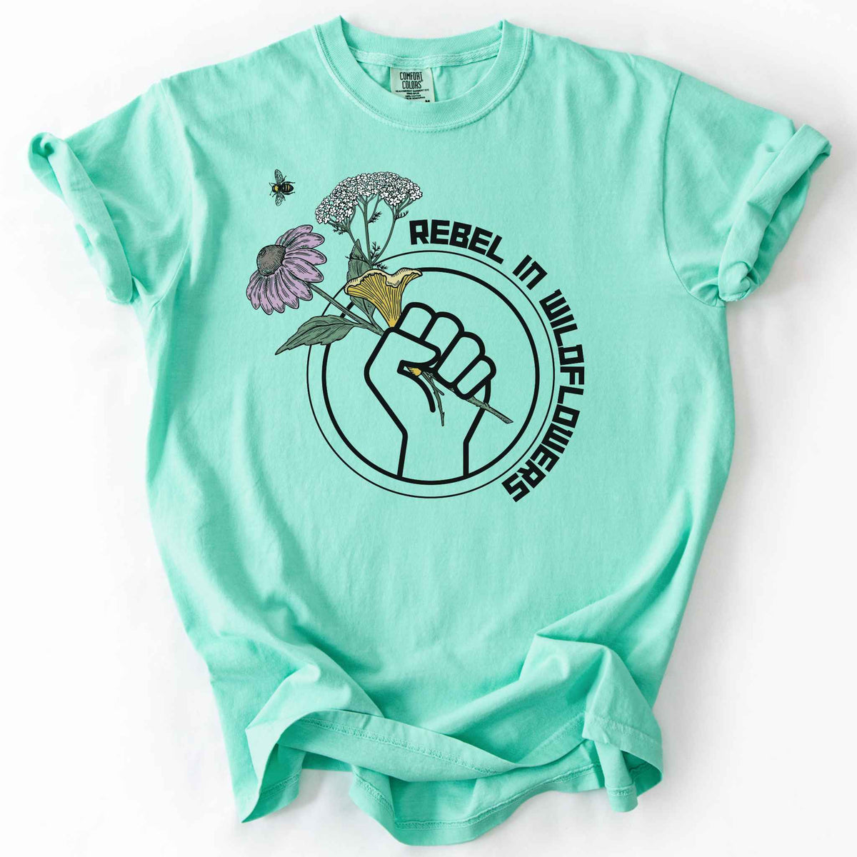 Island Reef "Rebel in Wildflowers" t-shirt on Comfort Colors tee, featuring a raised fist holding coneflower, yarrow, and a mushroom with a bumblebee in a circular badge design. Gardener and native plant gift shirt for wildflower and pollinator conservation fans.