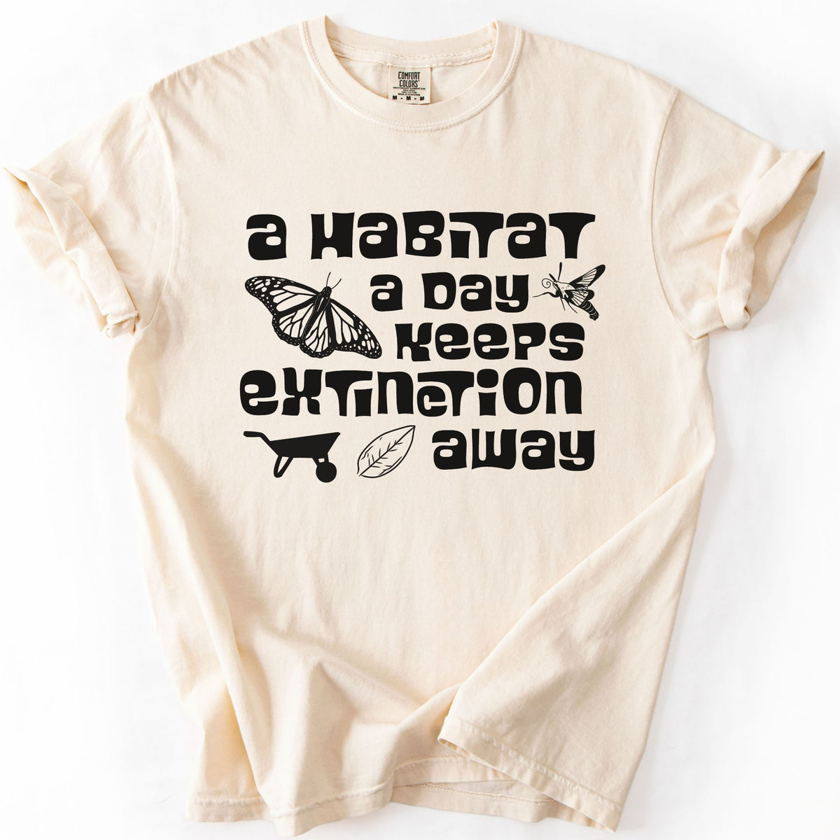 Ivory "A Habitat a Day Keeps Extinction Away" t-shirt on Comfort Colors tee, featuring a monarch butterfly, clearwing moth, wheelbarrow, and leaf icon with bold black lettering. Gardener and conservation gift shirt for native plant and pollinator enthusiasts.