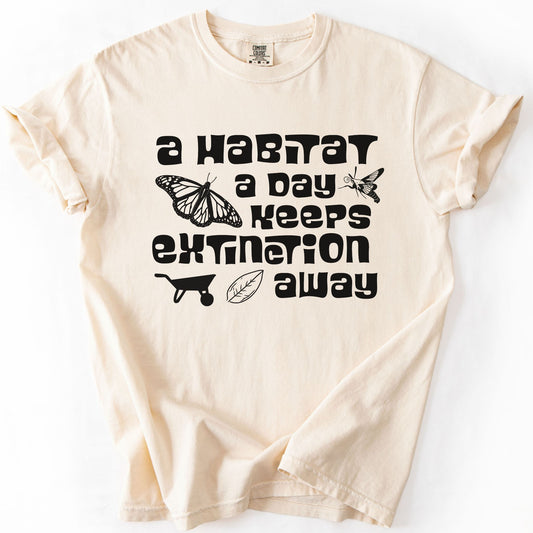 Ivory "A Habitat a Day Keeps Extinction Away" t-shirt on Comfort Colors tee, featuring a monarch butterfly, clearwing moth, wheelbarrow, and leaf icon with bold black lettering. Gardener and conservation gift shirt for native plant and pollinator enthusiasts.