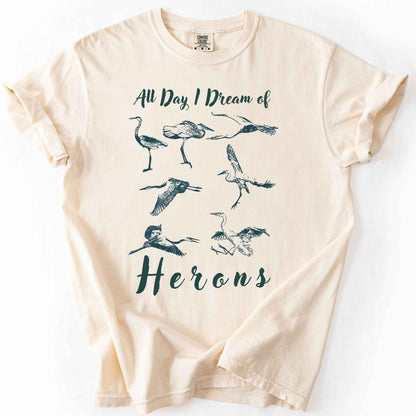 Ivory "All Day I Dream of Herons" t-shirt on Comfort Colors tee, featuring seven herons in various poses including standing, flying, and hunting. Birdwatcher and wading bird lover gift shirt for wildlife and nature enthusiasts.