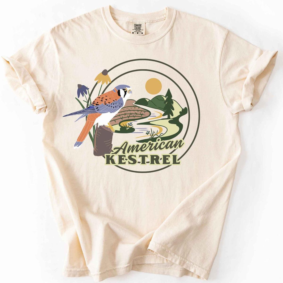 Ivory "American Kestrel" t-shirt on Comfort Colors tee, featuring a colorful illustrated kestrel perched among wildflowers with a farmland and river landscape in a circular badge design. Birdwatcher and raptor lover gift shirt for wildlife and nature enthusiasts.