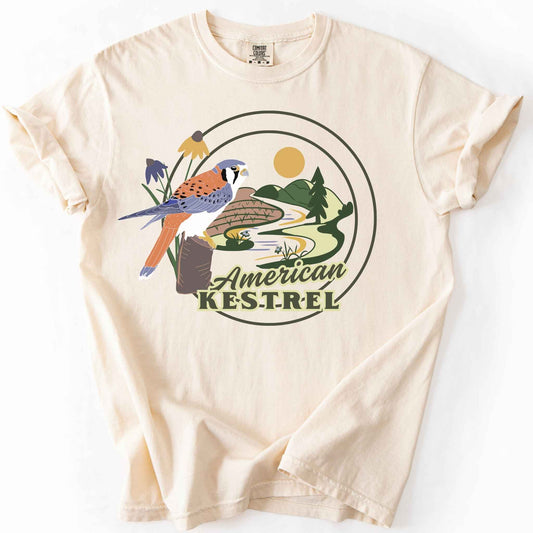 Ivory "American Kestrel" t-shirt on Comfort Colors tee, featuring a colorful illustrated kestrel perched among wildflowers with a farmland and river landscape in a circular badge design. Birdwatcher and raptor lover gift shirt for wildlife and nature enthusiasts.