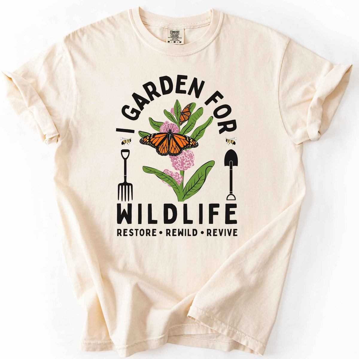 Ivory "I Garden for Wildlife, Restore, Rewild, Revive" t-shirt on Comfort Colors tee, featuring monarch butterflies and bees on milkweed flowers flanked by garden tools in orange and gold tones. Gardener and native plant gift shirt for pollinator conservation fans.