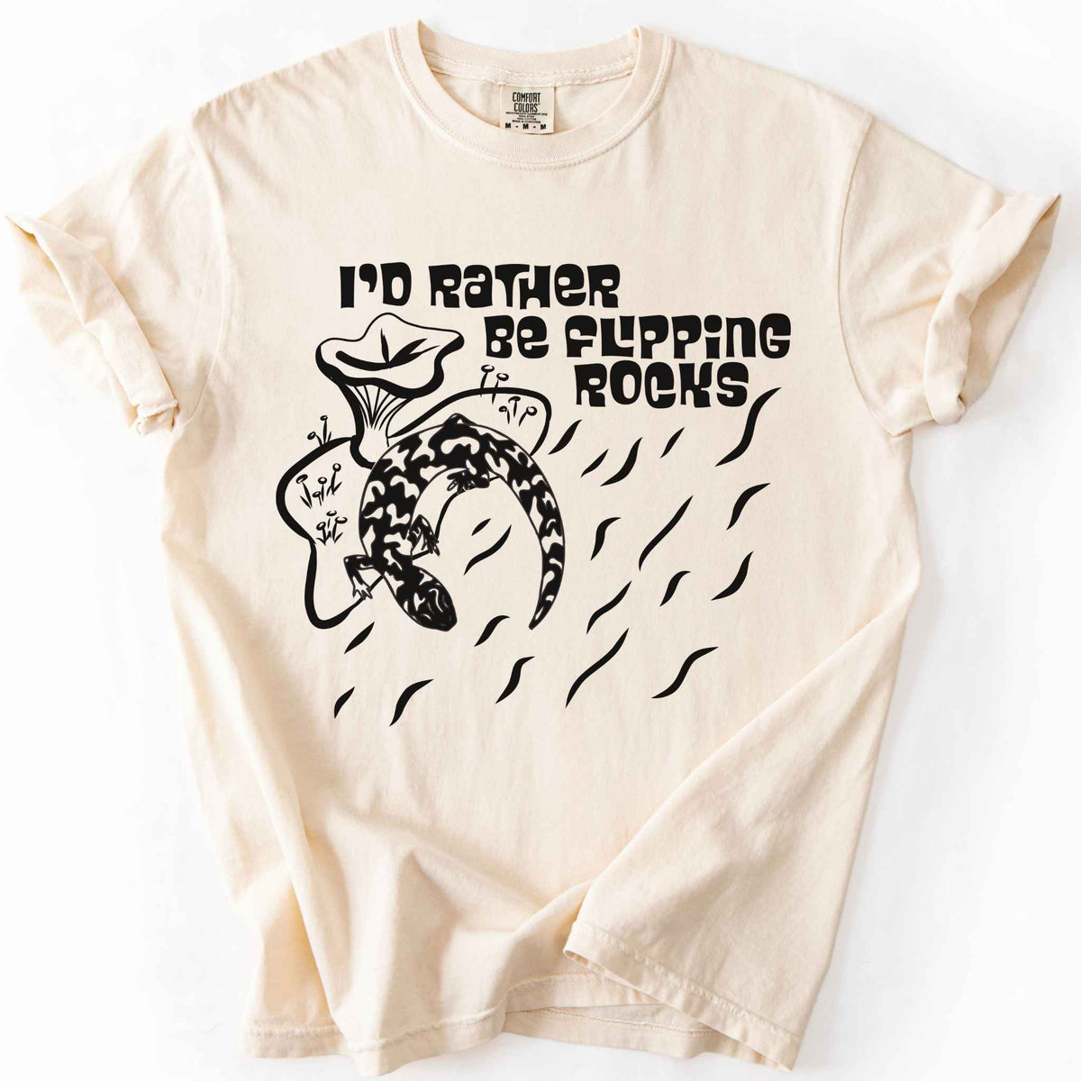 Ivory "I'd Rather Be Flipping Rocks" t-shirt on Comfort Colors tee, featuring a bold graphic of a curled salamander with mushrooms in a groovy retro font. Funny herpetology gift shirt for salamander hunters and amphibian enthusiasts.