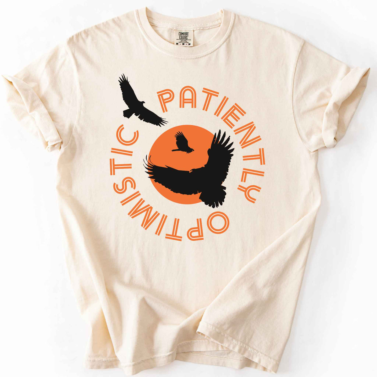 Ivory "Patiently optimistic turkey vulture" t-shirt on comfort colors tee. Silhouette turkey vulture graphics against an orange sun surrounded by curved text. Bird lover and wildlife enthusiast gift. 