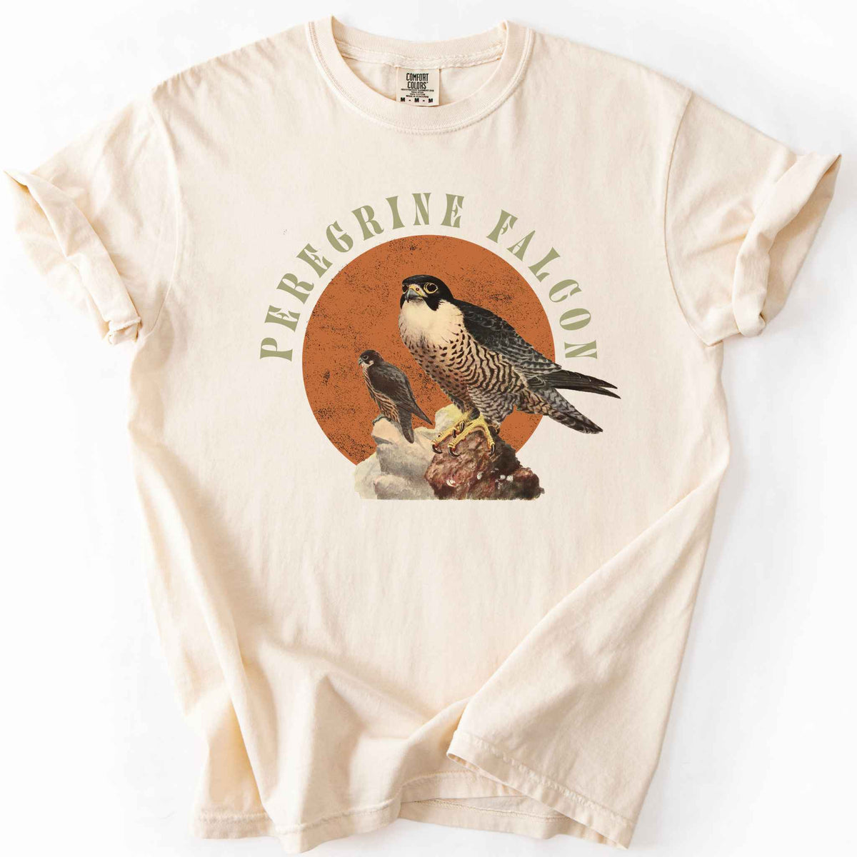 Ivory "Peregrine Falcon" t-shirt on Comfort Colors tee, featuring vintage style falcon illustration perched on a rock against an orange sun backdrop, with arched cream text. Birdwatcher and raptor lover gift shirt for falconry and wildlife fans.
