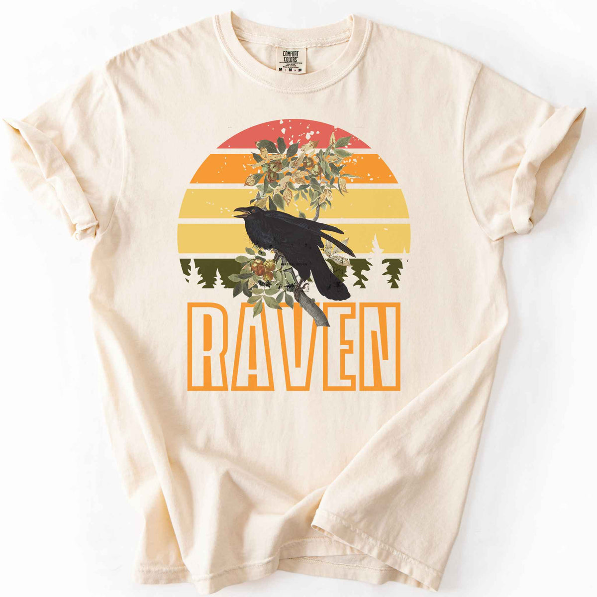 Ivory "Raven" t-shirt on Comfort Colors tee, featuring a raven perched on a branch against a retro sunset sunburst design with bold orange text. Bird lover and wildlife gift shirt for corvid and nature enthusiasts.