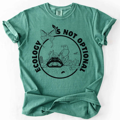 Light Green "Ecology Is Not Optional" t-shirt on Comfort Colors tee, featuring a black circle badge with a box turtle, bird, luna moth, frog, and mushrooms in an ecosystem scene. Nature lover and biology gift shirt for ecologists and conservation enthusiasts.