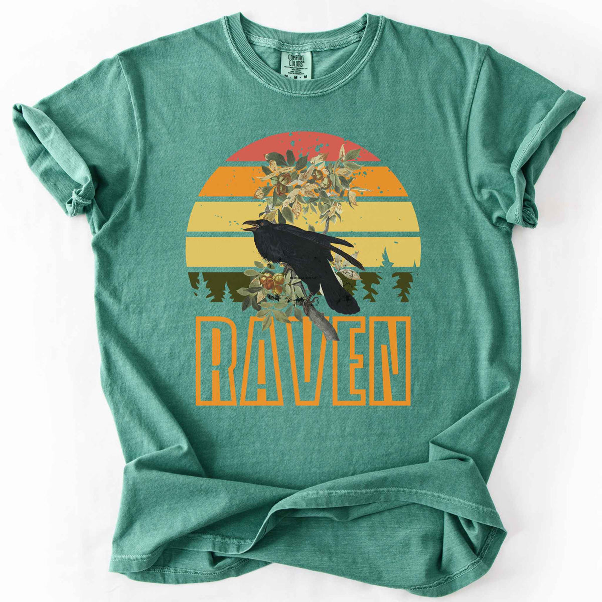Light Green "Raven" t-shirt on Comfort Colors tee, featuring a raven perched on a branch against a retro sunset sunburst design with bold orange text. Bird lover and wildlife gift shirt for corvid and nature enthusiasts.